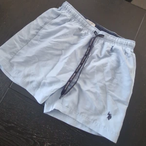 Ljusblå badshorts  - Ljusblå shorts från U.S. Polo Assn med elastisk midja och snörning med logga. Klassisk design med broderad logotyp på benet och meshfoder på insidan. Perfekta för sommardagar eller stranden.