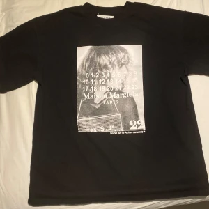Maison Margiela svart t-shirt med print - Svart t-shirt från Maison Margiela med stort grafiskt print framtill i svartvitt. Klassisk rund hals och korta ärmar. Tillverkad i mjuk bomull, perfekt för dig som gillar statement-plagg och streetstyle.