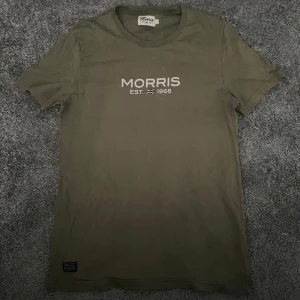 Olivgrön t-shirt från Morris - Olivgrön t-shirt från Morris med tryck 'MORRIS EST. 1968' och liten brittisk flagga på bröstet. Klassisk rund hals och korta ärmar. Skön och enkel modell i bomull. Hör av er vid frågor eller funderingar 