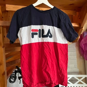 FILA t-shirt i rött, vitt och marinblått - Snygg t-shirt från FILA med breda blockränder i marinblått, vitt och rött. Stor logga framtill, klassisk rund hals och korta ärmar. Skön bomullskänsla och perfekt för en ”sportig” look. Den används aldrig så tänker den kanske kan komma till hands för någon att använda.