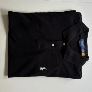 Svart pikétröja från Ralph Lauren - Klassisk svart pikétröja från Ralph Lauren med vit broderad logga på bröstet. Tröjan har korta ärmar, krage och två knappar framtill. Bara haft på få tal gånger