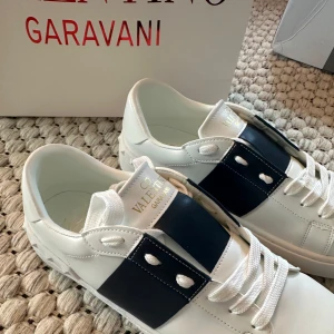 Valentino Garavani Open sneakers vit/blå - Snygga Valentino Garavani Open sneakers i vitt skinn med bred marinblå rem över mitten. Klassisk rund tå, vita snören och coola nitar på sulan. Perfekta för dig som vill sticka ut med exklusiva detaljer och stilren design.