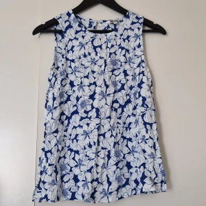 Blommig blå och vit ärmlös blus - Superfin ärmlös blus från Indiska med blå botten och vitt blommönster. Toppen har rund halsringning och knapp i nacken. Perfekt för dig som gillar en fräsch och somrig look med mycket mönster.