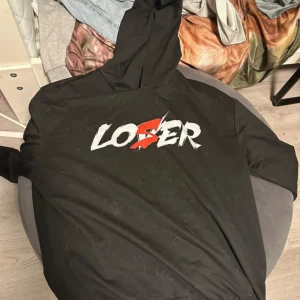 Svart hoodie med tryck 'LOSER' - Svart hoodie med stort tryck på bröstet där det står 'LOSER' i vitt och rött. Tröjan har huva och långa ärmar, perfekt för en avslappnad streetstyle-look. Materialet känns mjukt och bekvämt, och trycket ger en edgy vibe. Har även matchande mjukisbyxor till