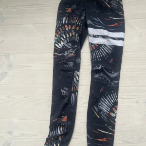 Mönstrade träningstights från Stronger XS - Snygga svarta träningstights från Stronger i storlek XS med färgglatt abstrakt mönster och två vita ränder på ena benet. Tillverkade i återvunnen polyester och elastan för en stretchig och bekväm passform. Perfekta för gymmet eller löprundan.