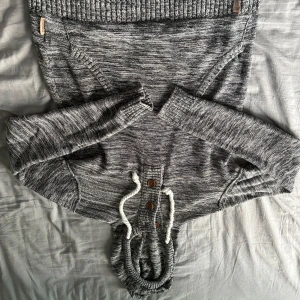 Gråmelerad stickad hoodie med knappar storlek M - Mysig gråmelerad stickad hoodie med långa ärmar, huva och vita snören. Tröjan har tre bruna knappar framtill och ribbade muddar vid ärmslut och nederkant. Perfekt för kyliga dagar och chill vibes.