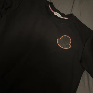 Svart Moncler sweatshirt med logga - Svart sweatshirt från Moncler med stor logga på bröstet och mindre logga på ärmen. Klassisk rund halsringning med ribbad kant och snygg detalj med rödvit rand på insidan av kragen. Perfekt för dig som gillar stilrena och exklusiva plagg.