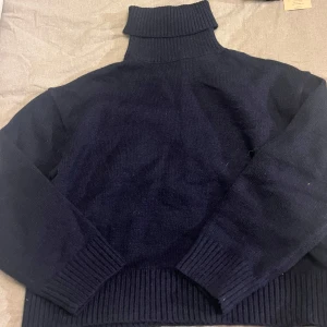 Mörkblå stickad polotröja - Mysig mörkblå stickad polotröja med hög krage och långa ärmar. Tröjan har breda ribbade muddar vid ärmslut och nederkant, perfekt för kyliga dagar. Enkel och stilren design som funkar till många outfits.