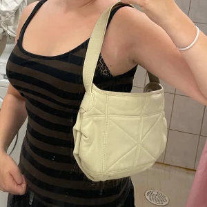 Beige axelväska från White Flirt - Säljer en off white axelväska från White Flirt. Väskan har ett quiltat mönster på utsidan och ett blommigt innerfoder i ljusa färger. Den har bred axelrem och dragkedja på insidan, perfekt för att bära med sig det viktigaste.