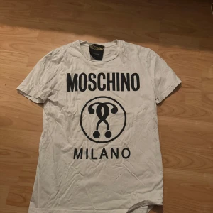 Vit Moschino Milano t-shirt - Vit t-shirt från Moschino med stor svart logga och Milano-tryck på bröstet. Klassisk rund halsringning och korta ärmar. T-shirten är i mjuk bomull och har en clean, stilren vibe som passar perfekt till jeans eller shorts.