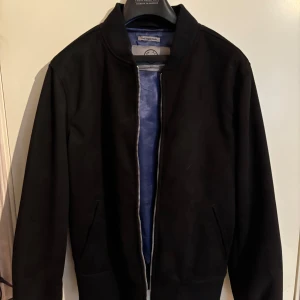 Svart bomberjacka från Human Scales - Stilren svart bomberjacka från Human Scales i vegansk mocka. Jackan har klassisk passform, ribbade muddar och silverfärgad dragkedja. Fodret är blått och ger en snygg kontrast. Perfekt för dig som vill ha en clean look med modern touch.