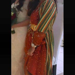 Färgstark kurdisk klänning med guldiga detaljer - Säljer en unik kurdisk klänning i orange med pärlbroderier och guldigt bälte. Ärmarna är långärmade med färgglada ränder i grönt, gult, vitt och rött. Klänningen har en traditionell look och är perfekt för dig som vill sticka ut med färg. Storlekarna Xs, S och M✨