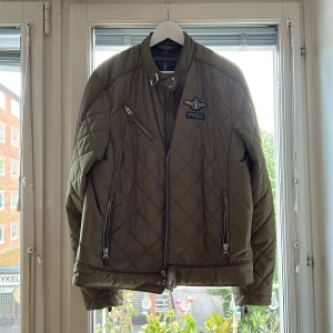 Olivgrön quiltad jacka från Morris - Säljer en olivgrön quiltad jacka från Morris Gent's med snyggt emblem på bröstet. Jackan har dragkedjor på fickor och bröst, hög krage med spänne och coola detaljer. Perfekt för dig som gillar klassisk men sportig stil.