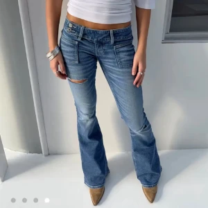 Blå bootcut jeans med slitningar - Snygga blå bootcut jeans med låg midja och slitningar på låret och knät. Jeansen har framfickor med lock och dubbla knappar i midjan. Perfekta för en avslappnad och trendig look.