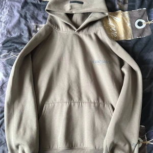 Beige Essentials Fear of God hoodie - Säljer en beige hoodie från Essentials Fear of God i storlek L. Hoodien har en klassisk känguruficka, huva och diskret logga på bröstet. Mjuk insida och relaxed fit gör den perfekt för chill dagar. Stilren och enkel att matcha med det mesta.