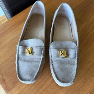 Beige mockaloafers från Lauren Ralph Lauren - Stilrena loafers i beige mocka från Lauren Ralph Lauren med guldfärgad RL-detalj framtill. Skorna har platt sula med gummidetaljer undertill för extra grepp och en klassisk, tidlös design. Perfekta för dig som gillar en clean och sofistikerad look.