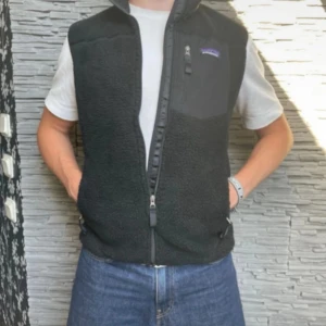 Svart fleeceväst från Patagonia - Svart fleeceväst från Patagonia med hög krage och hel dragkedja framtill. Västen har två sidofickor med dragkedja och en bröstficka med logga. Mjuk och varm, perfekt för lager-på-lager-stil under sommarkvällar eller kyliga dagar. Nypris 1400 kr, denna är så gott som ny! Fraktar oftast samma dag som köp😄