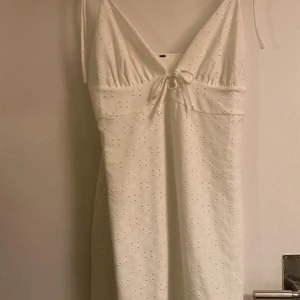 Vit somrig klänning med knyt - Superfin vit kortklänning med broderade hålmönster och smala axelband. V-ringad framtill med söt knytning under bysten. Perfekt för varma dagar och har en lätt och luftig känsla.