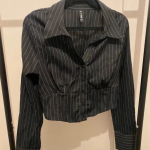 Svart croppad skjorta från FB Sister - Svart croppad skjorta från FB Sister med vita tunna ränder. Skjortan har lång ärm, klassisk krage och knäppning framtill. Snygg passform med markerad midja och elastisk rygg. Perfekt för dig som gillar en trendig och stilren look. Extra mitten knappen är sönder. Knappt använd