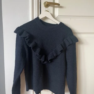 Mörkblå stickad volangtröja Gina Tricot - Supersöt mörkblå stickad tröja från Gina Tricot med volangdetaljer som går snett över framsidan och axlarna. Tröjan har lång ärm, rund halsringning och ett diskret vågigt mönster i stickningen. Perfekt för dig som gillar en trendig och mysig look.