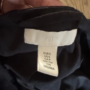 Svart långklänning från H&M - Svart långklänning från H&M med korta puffärmar och fina spetsdetaljer vid ärmslut och midja. Klänningen har en rund halsringning och en elegant, flowy siluett som ger en stilren look.