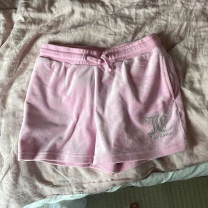 Rosa Juicy Couture shorts med snörning - Supermjuka rosa shorts från Juicy Couture i velour med snörning i midjan och silverbroderad logga framtill. Perfekta för chill dagar eller när du vill ha en ikonisk Y2K-look. Klassisk loose fit och bekvämt resårband i midjan. Supermjuk rosa zip hodde med en silvrig dragkedja 