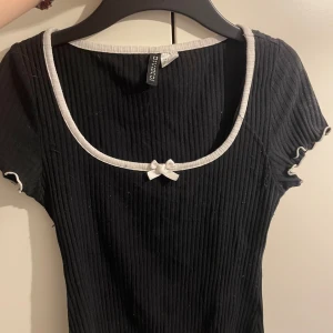 Svart ribbad topp med rosett H&M S - Supersöt svart ribbad topp från H&M med vit kant runt halsringningen och ärmsluten. Söt liten rosett framtill och lätt croppad passform. Perfekt för dig som gillar en enkel men gullig stil.