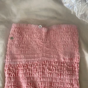 Rosa axelbandslös kortklänning - Supersöt rosa kortklänning med smockad överdel och volangig kjol i broderad bomull🩷. Axelbandslös modell som sitter tajt upptill och har två lager volanger nertill med fina utskurna detaljer. Perfekt för sommarens alla tillfällen!🏖️ Den är ALDRIG använd!