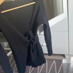 Svart ribbad omlottkofta med knytband - Svart långärmad ribbstickad omlottkofta med knytband i midjan. Koftan har en djup v-ringning och är gjord i ett mjukt, stickat material som passar perfekt för kyligare dagar. Enkel att styla och supermysig att bära.