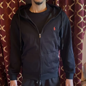 Svart hoodie från Polo Ralph Lauren - Svart hoodie från Polo Ralph Lauren med dragkedja, huva med snörning och klassisk röd broderad logga på bröstet. Tröjan har känguruficka framtill och är tillverkad i mjuk bomull. Perfekt för en chill och stilren look. Helt ny, säljer den då den var i fel storlek. Den är storlek S