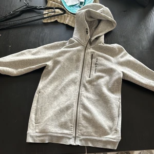 Grå hoodiejacka från Sail Racing - Snygg ljusgrå hoodiejacka från Sail Racing med dragkedja hela vägen, bröstficka med dragkedja och broderad logga. Mjuk fleece på insidan och huva med logotyp. Perfekt för dig som gillar sportig och clean stil.