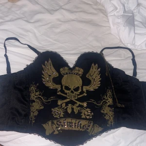 Svart korsett med dödskalletryck - Cool svart korsett med justerbara axelband och guldfärgat dödskalletryck med vingar och krona framtill. Dekorerad med små nitar och spetskant. Hakknäppning bak och detaljerade mönster på sidorna. Perfekt för dig som vill sticka ut med edgy stil. Storlek s passar xs 