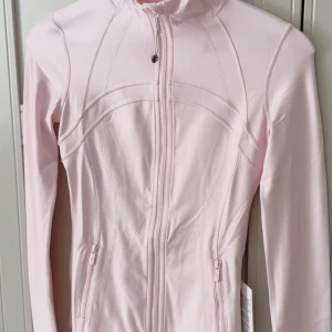 Ljusrosa define jacka från Lululemon - Helt ny supersnygg ljusrosa define jacka från Lululemon med dragkedja framtill och höga krage. Den har långa ärmar och två fickor med dragkedja. Perfekt för dig som gillar en clean girl stil.