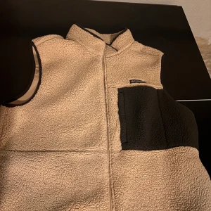  fleeceväst från Columbia - Mysig beige fleeceväst från Columbia med hög krage och svart bröstficka. Västen har dragkedja framtill och är perfekt för lager-på-lager. Fleeceytan ger en skön och avslappnad look.