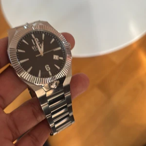Versus Versace silverfärgad klocka - Stilren klocka från Versus Versace med svart urtavla, datumvisning och markerade index. Boetten och länken är i blankt och borstat silverfärgat stål. Klockan har en räfflad bezel och klassisk länkarmband. Perfekt för dig som gillar en lyxig och modern look. Pris kan diskuteras vid snabb affär 