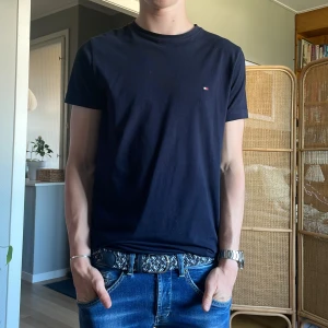 Mörkblå t-shirt från Tommy Hilfiger - Snygg mörkblå t-shirt från Tommy Hilfiger med liten broderad logga på bröstet. Klassisk passform och rund halsringning. Tillverkad i mjuk bomull som känns skön mot huden. Perfekt för en clean och stilren look.