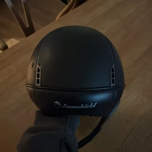 Ridhjälm från Samshield - Snygg svart ridhjälm från Samshield med med solskärm. Den e i storlek S vilket motsvarar 52-56cm. Den har en löstagbar liner vilket är perfekt så att man kan tvätta den. Linern är i storlek S men eftersom att den är löstagbar gör det att byta storlek. Kommer med hjälmskydd!!!