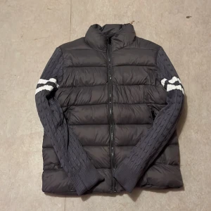 Moncler pufferjacka med stickade ärmar - Snygg mörkgrå pufferjacka från Moncler med stickade ärmar i samma färg och vita ränder. Jackan har dragkedja framtill och Moncler-logga på ena ärmen. Perfekt för dig som vill ha en unik och sportig look under höst och vinter.pris kan diskuteras 