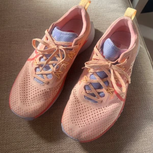 Nike React löparskor i persika och lila - Nike React löparskor i snygg persikofärg med lila mesh på plösen och detaljer i orange och rött. Skorna har ventilerande ovandel, mönstrade snören och grov sula för bra grepp. Perfekta för dig som vill sticka ut på löprundan!