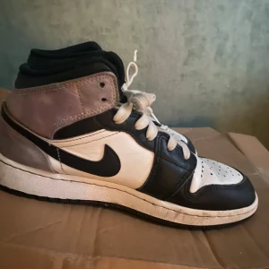 Nike Air Jordan 1 svart/vit/lila  - Säljer ett par Nike Air Jordan 1 med svart och vit läderöverdel, lila detaljer vid hälen och klassisk svart swoosh. Skorna har högt skaft, perforerad tå och vita snören. Perfekt för dig som gillar streetwear och vill sticka ut med en ikonisk sneaker.