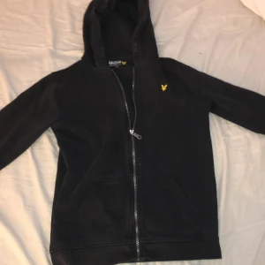 Svart hoodie från Lyle & Scott Junior - Svart hoodie från Lyle & Scott Junior med dragkedja framtill och klassisk gul logga på bröstet. Tröjan har huva, känguruficka och är tillverkad i mjuk bomull. Perfekt för en chill och stilren look. Kan sänka pris!!!