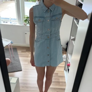 Ljusblå jeansklänning från Zara - Snygg ljusblå jeansklänning från Zara med knappar hela vägen framtill, två bröstfickor och råa ärmslut. Ärmlös modell med klassisk krage och cool denimkänsla. Perfekt för dig som gillar en avslappnad men trendig stil. Trendig nu i sommar. Säljer då den är för liten