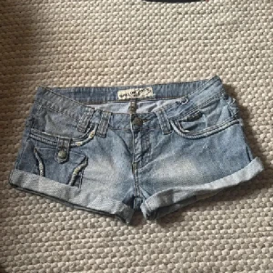 Gallop ljusblå jeansshorts med detaljer - Snygga ljusblå jeansshorts från Gallop med uppvikta ben och coola fickdetaljer med knappar och dekorativa sömmar. Klassisk femficksmodell och bälteshällor, perfekt till sommaren. Shortsen har låg midja och är gjorda i mjukt jeanstyg. midejemått ca 41 cm. Tryck ej köp nu