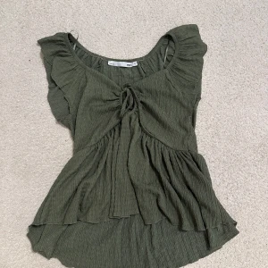 Grön volangblus med knyt från 157 - Supersöt grön blus från 157 med volangdetaljer och knyt framtill. Toppen har korta ärmar och en flowy, luftig passform med rynkade partier. Perfekt för dig som gillar en romantisk och chill stil.