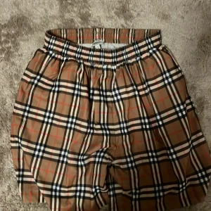 Rutig Burberry shorts XL - Snygga shorts från Burberry i klassiskt beige, svart, vit och röd rutigt mönster. De har elastisk midja med snörning och meshfoder på insidan. Perfekta för sommaren och ger en lyxig vibe till din outfit.