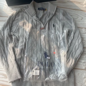 Grå kabelstickad tröja Polo Ralph Lauren - Kabelstickad grå tröja från Polo Ralph Lauren med halv dragkedja och klassisk krage. Tröjan har lång ärm, ribbade muddar och den ikoniska broderade loggan på bröstet. Perfekt för dig som gillar stilren och tidlös design. Finns ingen tvättlapp i 