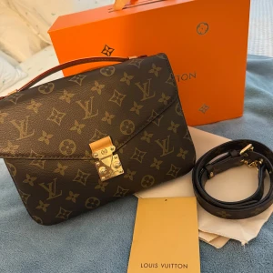 Louis Vuitton Pochette Métis - Säljer en klassisk Louis Vuitton handväska i brunt skinn med ikoniskt monogrammönster och guldfärgat lås. Väskan har både handtag och avtagbar axelrem, samt flera fack inuti för smart organisering. Perfekt för dig som vill ha en tidlös och lyxig accessoar.