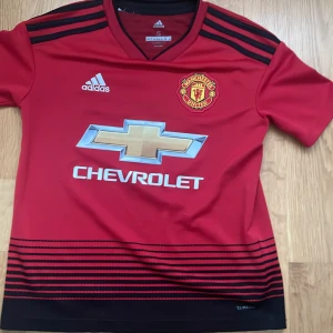 Manchester United Pogba tröja Adidas S - Manchester United matchtröja från Adidas med Pogba och nummer 6 på ryggen. Röd med svarta detaljer och ränder längst ner, klubbmärke och sponsortryck på bröstet. Tillverkad i lätt och ventilerande Climalite-material. Perfekt för dig som älskar fotboll och United.