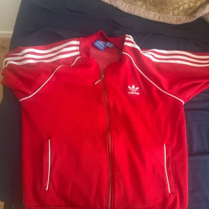 Röd Adidas track jacket med vita ränder - Riktigt snygg röd Adidas track jacket med klassiska vita ränder längs ärmarna och vit piping. Dragkedja framtill, två fickor och Adidas logga på bröstet. Perfekt för en sportig och retro vibe.