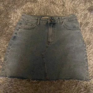 Grå jeanskjol med rå kant - Snygg grå jeanskjol från Bershka med klassisk femficksdesign och rå, fransig kant nertill. Kjolen har bälteshällor och dragkedja framtill. Perfekt för dig som gillar en avslappnad och trendig stil.